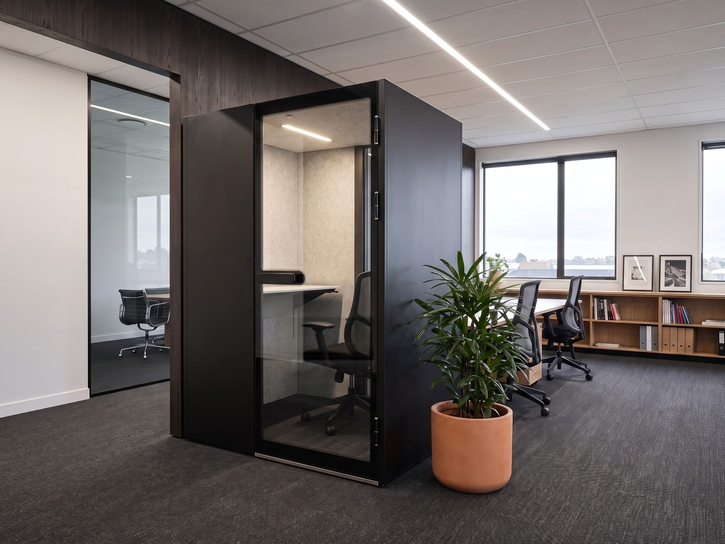 An MW-Pod against a dark timber wall in a quiet office, with a potted plant in a terracotta pot in the foreground and desks along the windows beyond