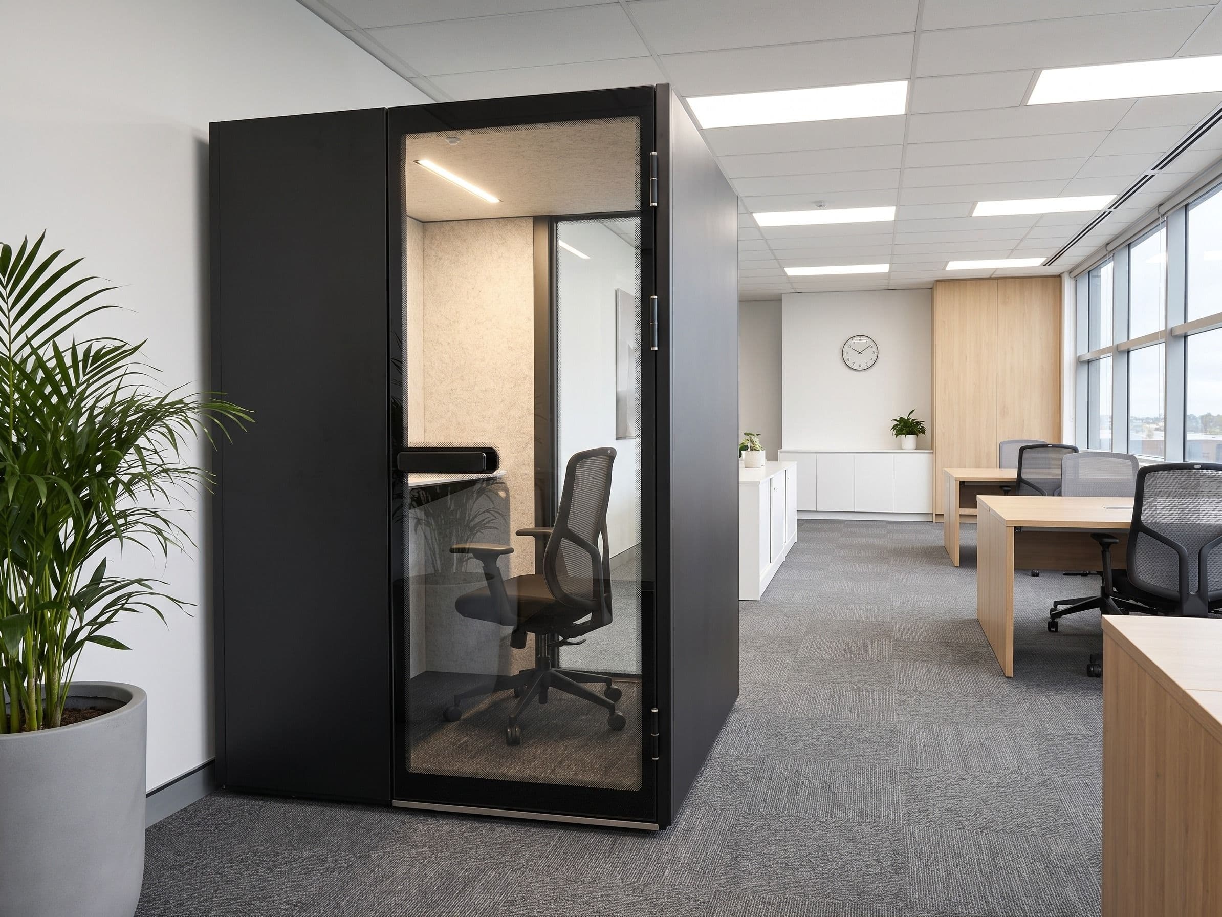 An MW-Pod against a white wall in a tidy open-plan office, with oak desks, a low white credenza and a wall clock beyond, and a potted palm in the foreground